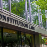 Constitution Cafe storefront in Washington, DC featuring dimensional lettering signage on a modern building facade with large windows, urban streetscape with trees, and pedestrian walkway